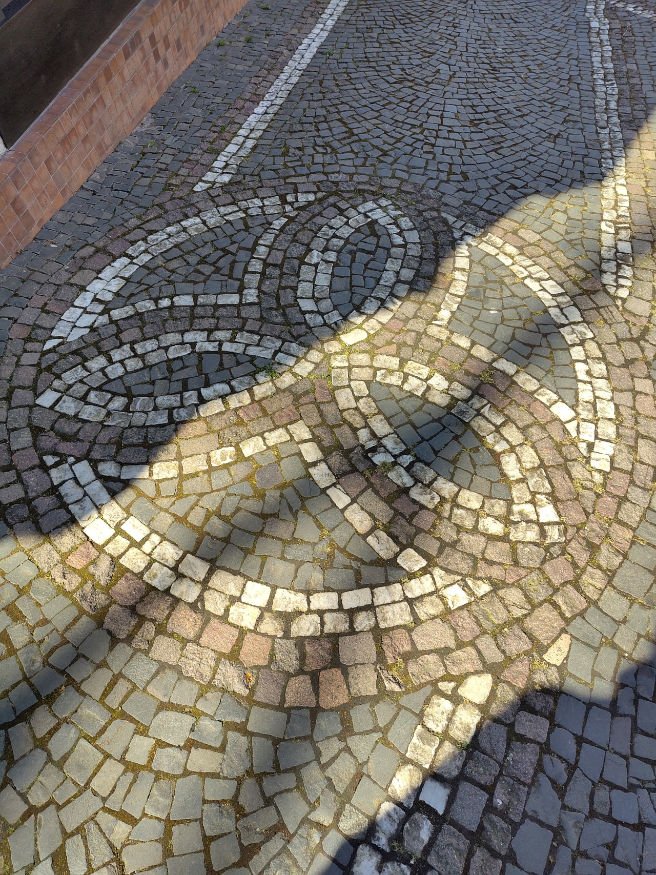 Ein gepflasterter Weg mit einem dekorativen Muster aus Steinen, das eine Spirale oder ein Labyrinth darstellt. Der Weg ist von einer Ziegelmauer und Bäumen gesäumt, und das Sonnenlicht wirft Schatten auf das Pflaster