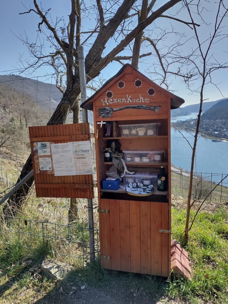 Ein kleines Holzhäuschen mit der Aufschrift 'Hexenküche' steht in einer ländlichen Umgebung mit Blick auf einen Fluss und Berge. Das Häuschen enthält verschiedene Gegenstände wie Flaschen, Krüge, und eine Puppe. Neben dem Häuschen befindet sich ein Schild mit Informationen und einem Zeitplan.