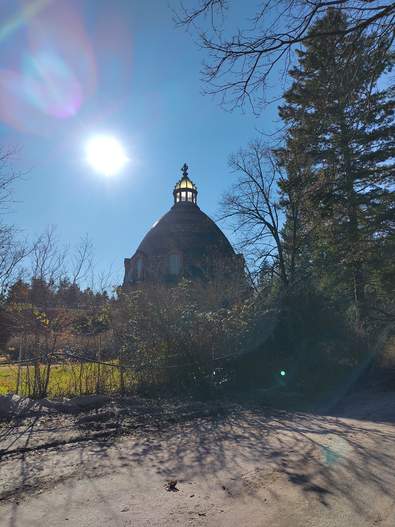 "Ein Gebäude mit einer Kuppel und einem Turm, das sich hinter Bäumen und Büschen befindet. Die Sonne scheint hell am Himmel und wirft Lichtflecken durch die Äste der Bäume. Im Vordergrund ist ein sandiger oder staubiger Weg zu sehen